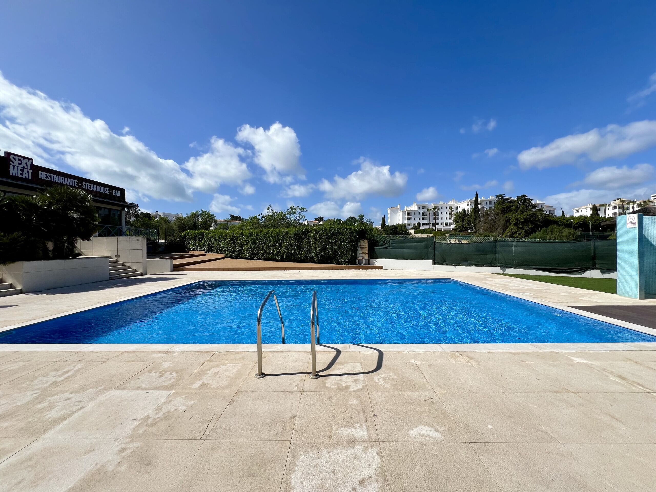 Ein Pool mit Metallleitern, umgeben von einer gefliesten Terrasse. Im Hintergrund ist ein Gebäude mit der Aufschrift "SEMP RESTAURANTE STEAKHOUSE BAR" zu sehen.