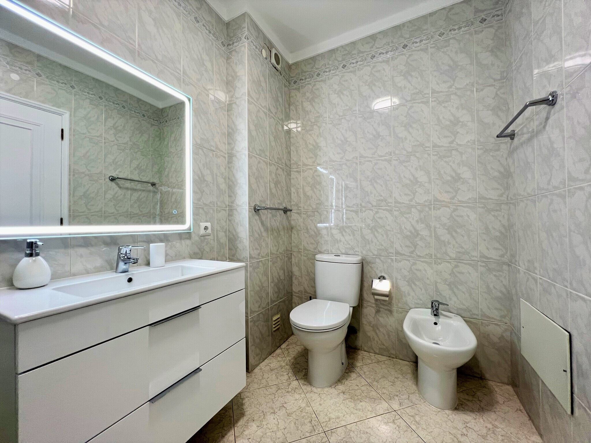 Et moderne badeværelse med et hvidt skab, toilet, bidet og et stort spejl.