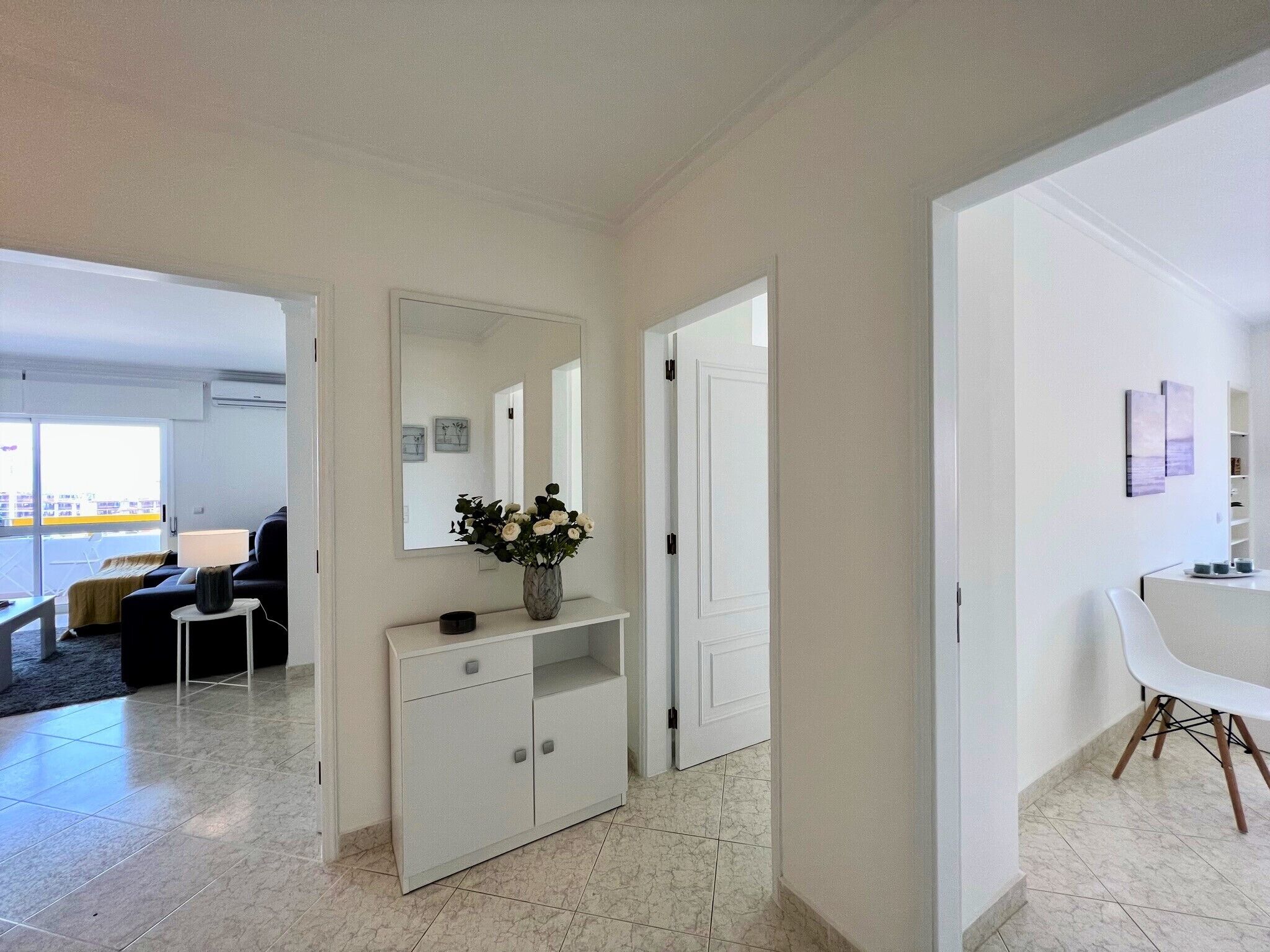 Uma sala de estar moderna com um sofá, uma mesa de centro e um armário branco com um vaso de flores. Há um espelho grande na parede e uma porta que dá para outro cômodo.