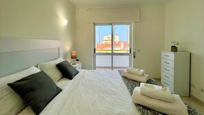 A hotel room with a bed, a nightstand, a dresser, and a view of buildings through a glass door. - Quarteira Panoramic View by Homing (Loulé)