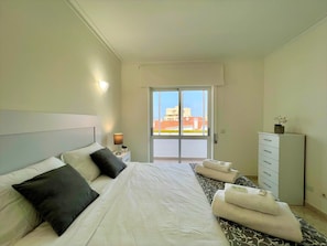 Ein Hotelzimmer mit Bett, Nachttisch, Kommode und Blick auf Gebäude durch eine Glastür.