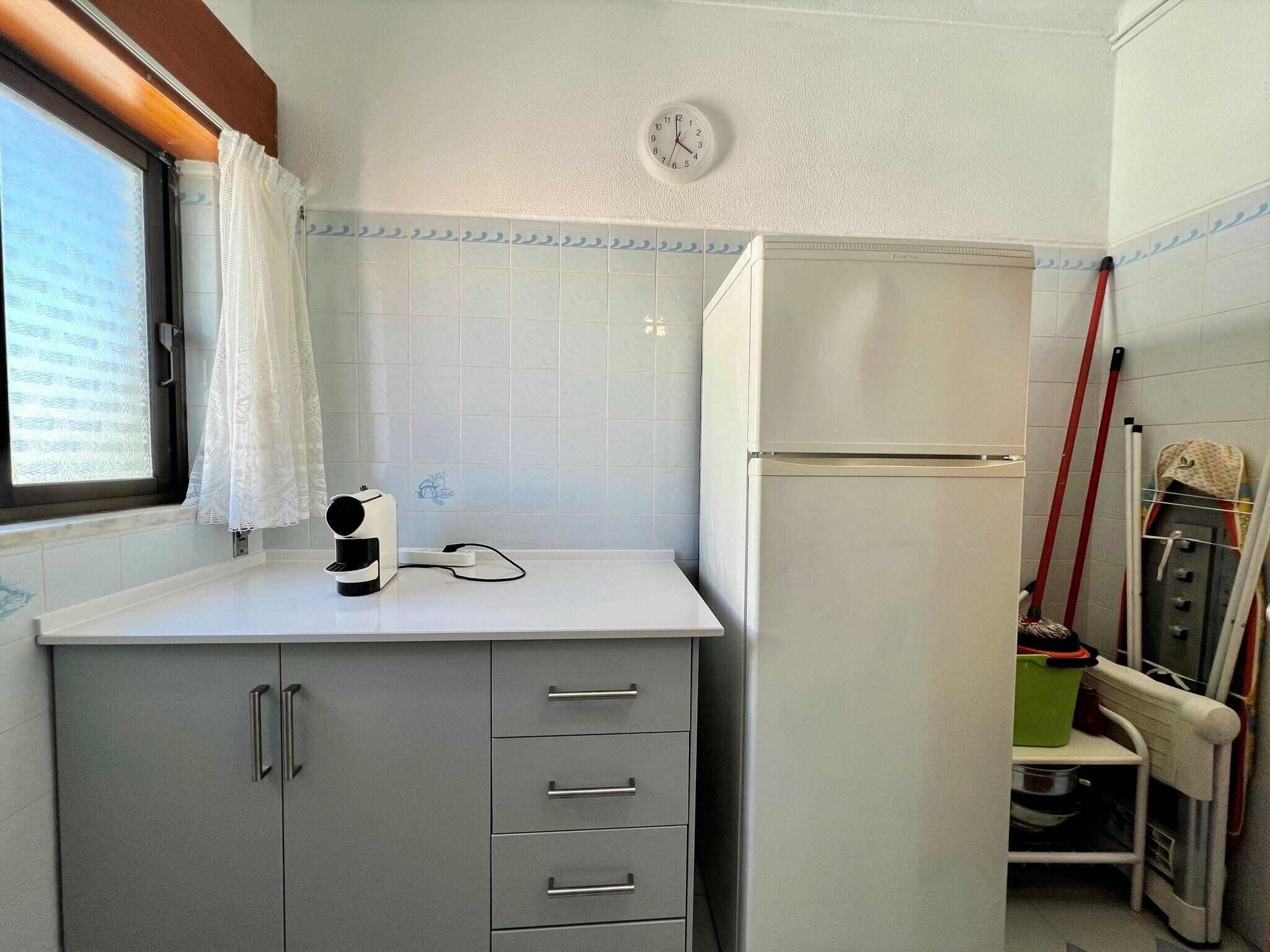 Una cocina compacta con un refrigerador blanco, un reloj en la pared y una ventana con cortina.