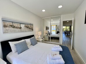 A modern hotel room with a large bed, a bedside table with a lamp, a painting on the wall, and a view of the dining area through a glass door. - Quarteira Stylish With Pool by Homing (Loulé)