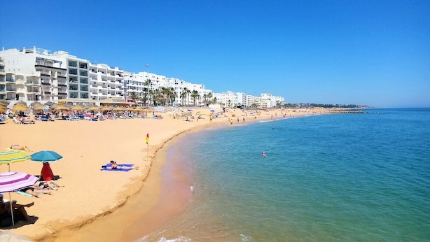 A beach with white buildings, clear blue water, and umbrellas. - Quarteira Stylish With Pool by Homing (Loulé)