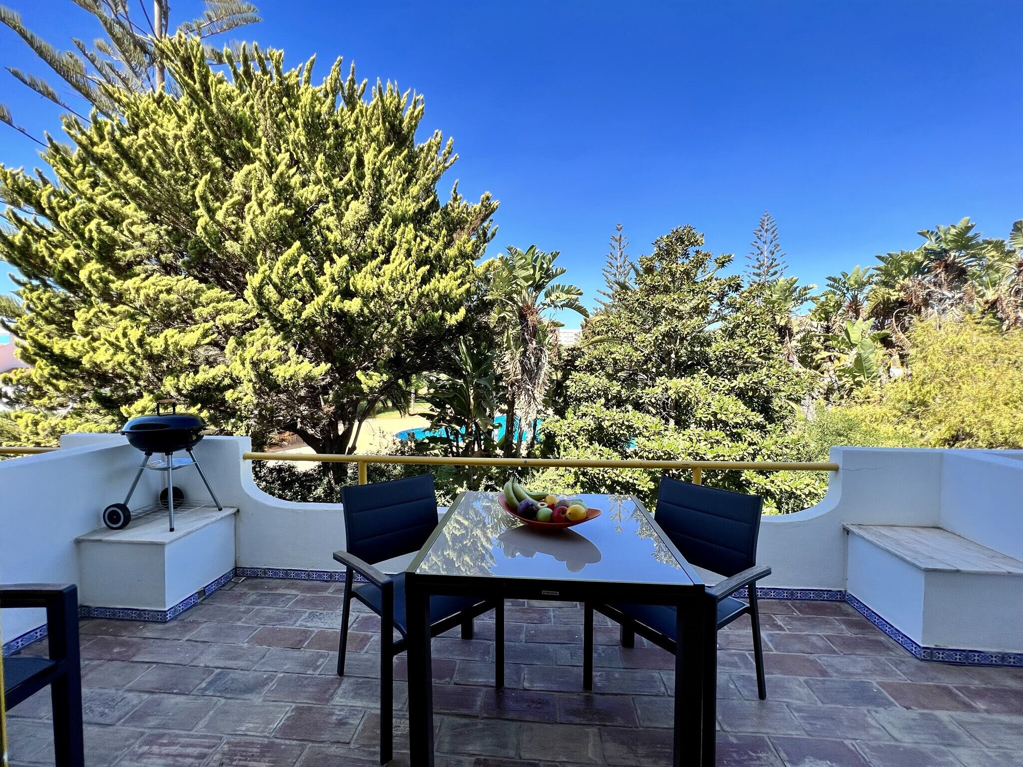 A patio with a table and chairs, a barbecue grill, and a view of a pool and trees.