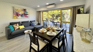 A modern living room with a dining area, a grey sofa with colorful cushions, a painting on the wall, and large windows offering a view of greenery. - Quarteira Stylish With Pool by Homing (Loulé)
