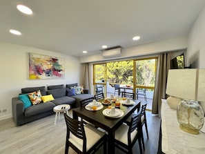 A modern living room with a dining area, a grey sofa with colorful cushions, a painting on the wall, and large windows offering a view of greenery. - Quarteira Stylish With Pool by Homing (Loulé)