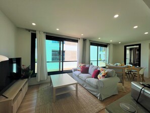 A modern living room with a sofa, coffee table, and a TV. There are large windows with curtains, a dining area with chairs, and a small kitchen area. - Faro Design 4 by Homing (Faro)