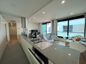 A modern kitchen with a built-in dishwasher, a coffee maker, and a sink. There is a dining area with a table and chairs, and a balcony with a view of the city. - Faro Design 4 by Homing (Faro)