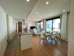 A modern kitchen with a central island, dining area with a table and chairs, and a large window with curtains. - Faro Design 4 by Homing (Faro)