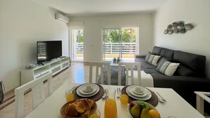 A modern living room with a flat-screen TV, a dark sofa, and a dining area with breakfast set on a white table. - Vilamoura Central 2 With Pool by Homing (Loulé)