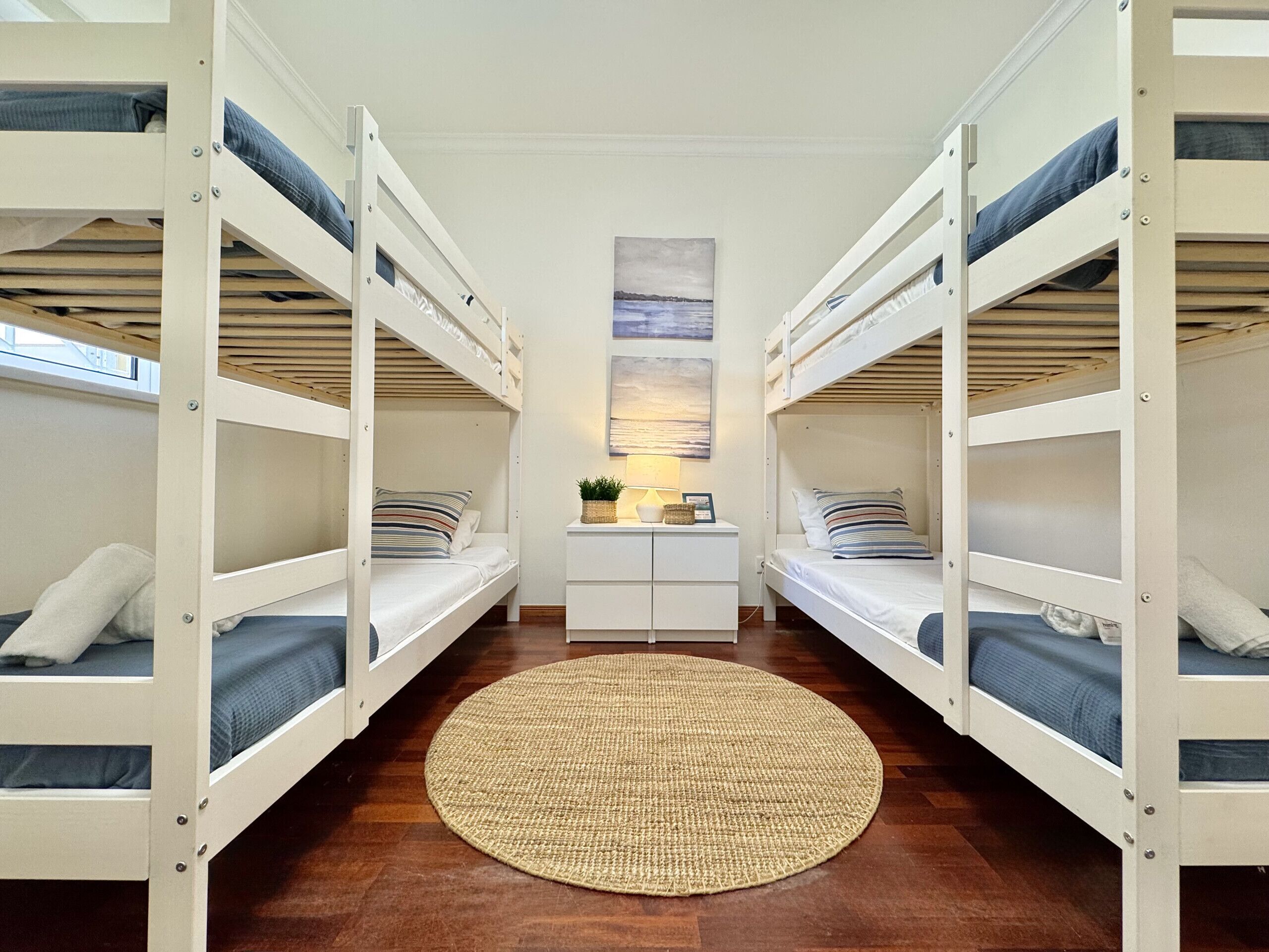 A room with bunk beds, a central white dresser, a woven rug, and a painting on the wall.