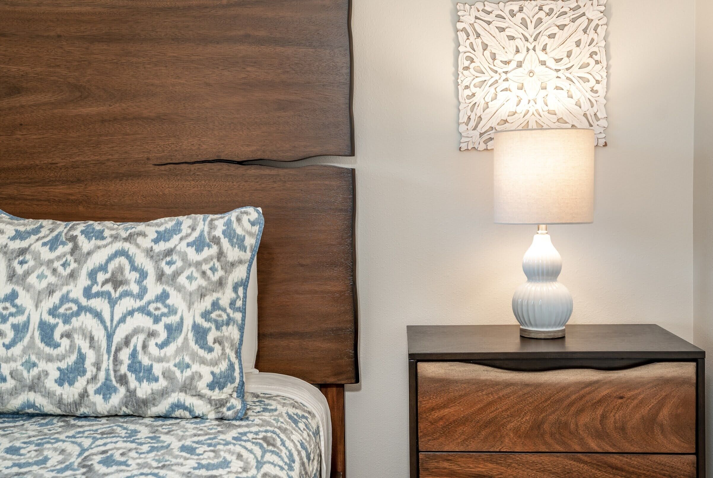 A bedroom with a wooden headboard, a patterned pillow, a bedside table with a lamp, and a decorative wall piece.