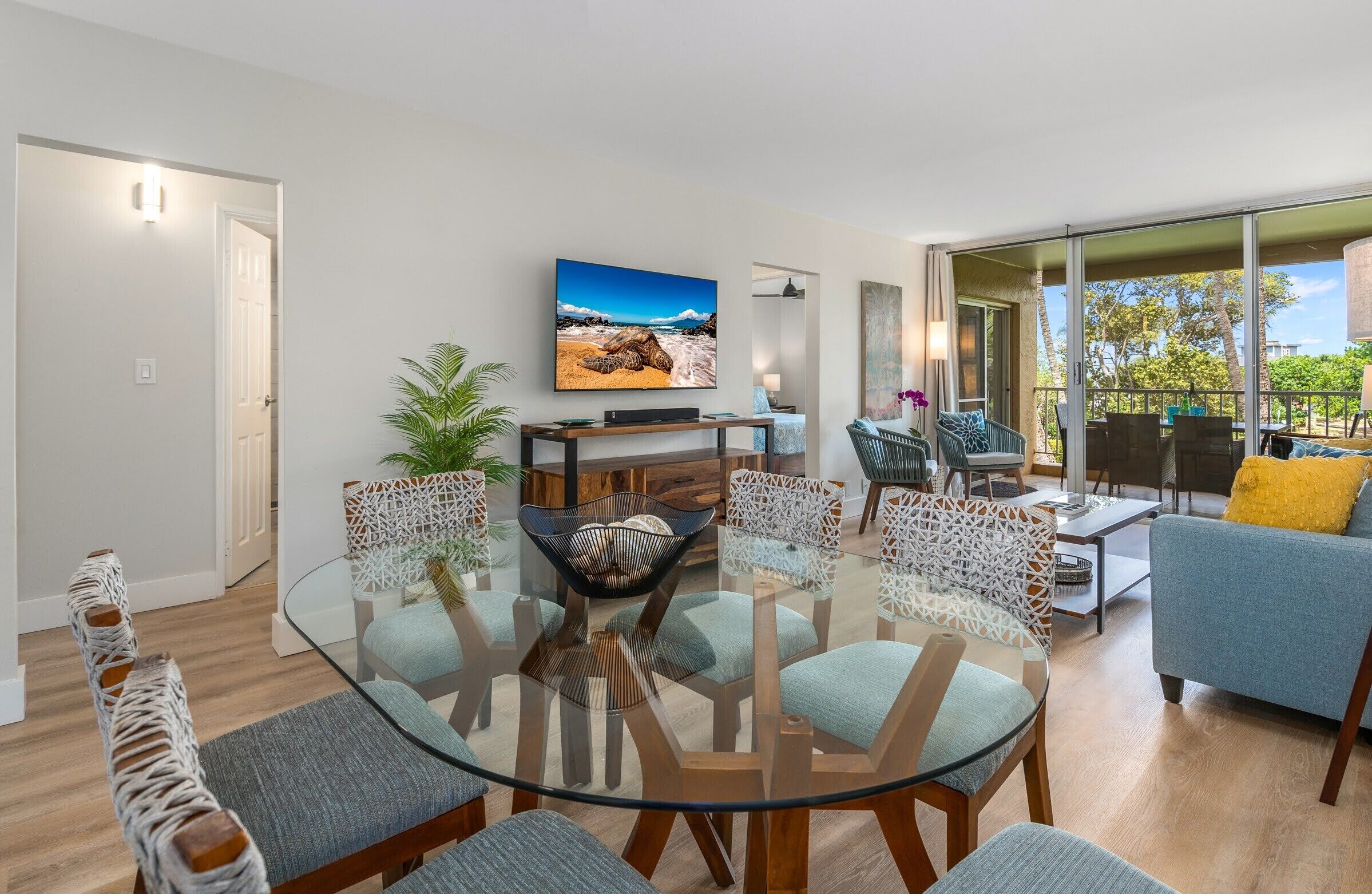 Un salon moderne avec une table basse en verre, des sièges confortables, un téléviseur à écran plat et un balcon aménagé avec du mobilier d’extérieur.