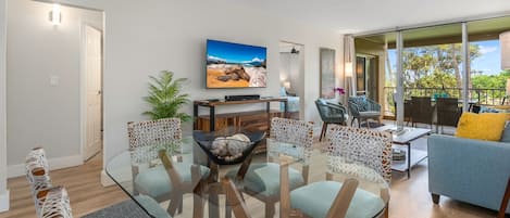 Un salon moderne avec une table basse en verre, des sièges confortables, un téléviseur à écran plat et un balcon aménagé avec du mobilier d’extérieur.