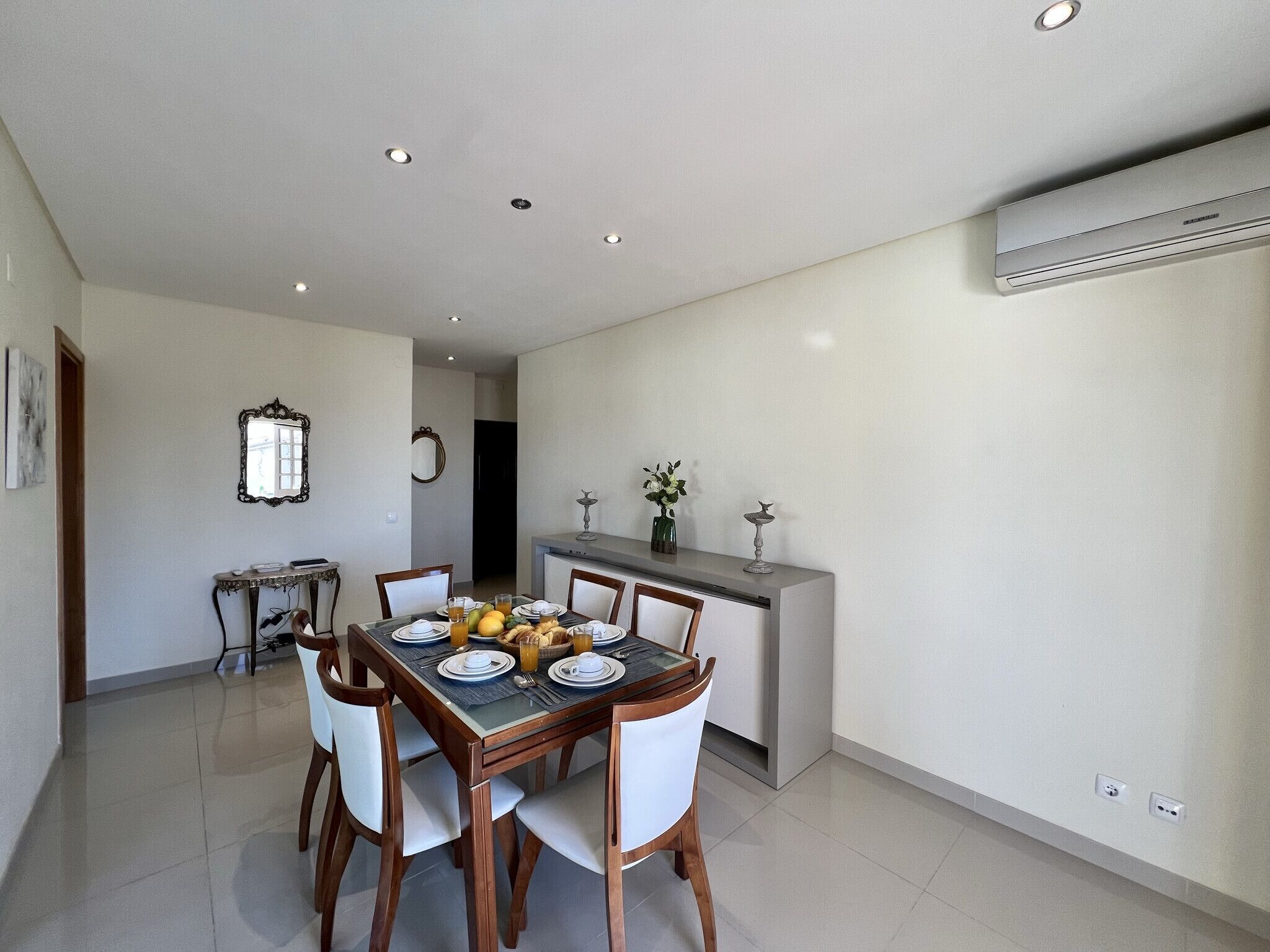 A dining area with a table set for four, chairs, a small side table, and a decorative mirror on the wall.