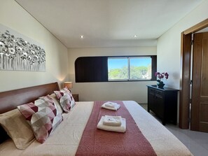 A bedroom with a bed, pillows, a bedside table with a lamp, a dresser, and a window with a view of trees. - Quarteira Classic With Pool by Homing (Loulé)