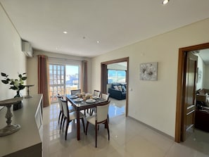 A dining area with a table set for four, a sofa in the background, and a city view through large windows. - Quarteira Classic With Pool by Homing (Loulé)