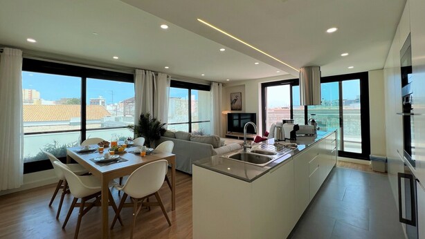 A modern kitchen with a central island, dining area with chairs, and large windows offering a city view. - Faro Design 2 by Homing (Faro)
