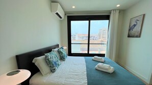 A hotel room with a bed, a bedside lamp, a window with a view of buildings, and an air conditioning unit. - Faro Design 2 by Homing (Faro)