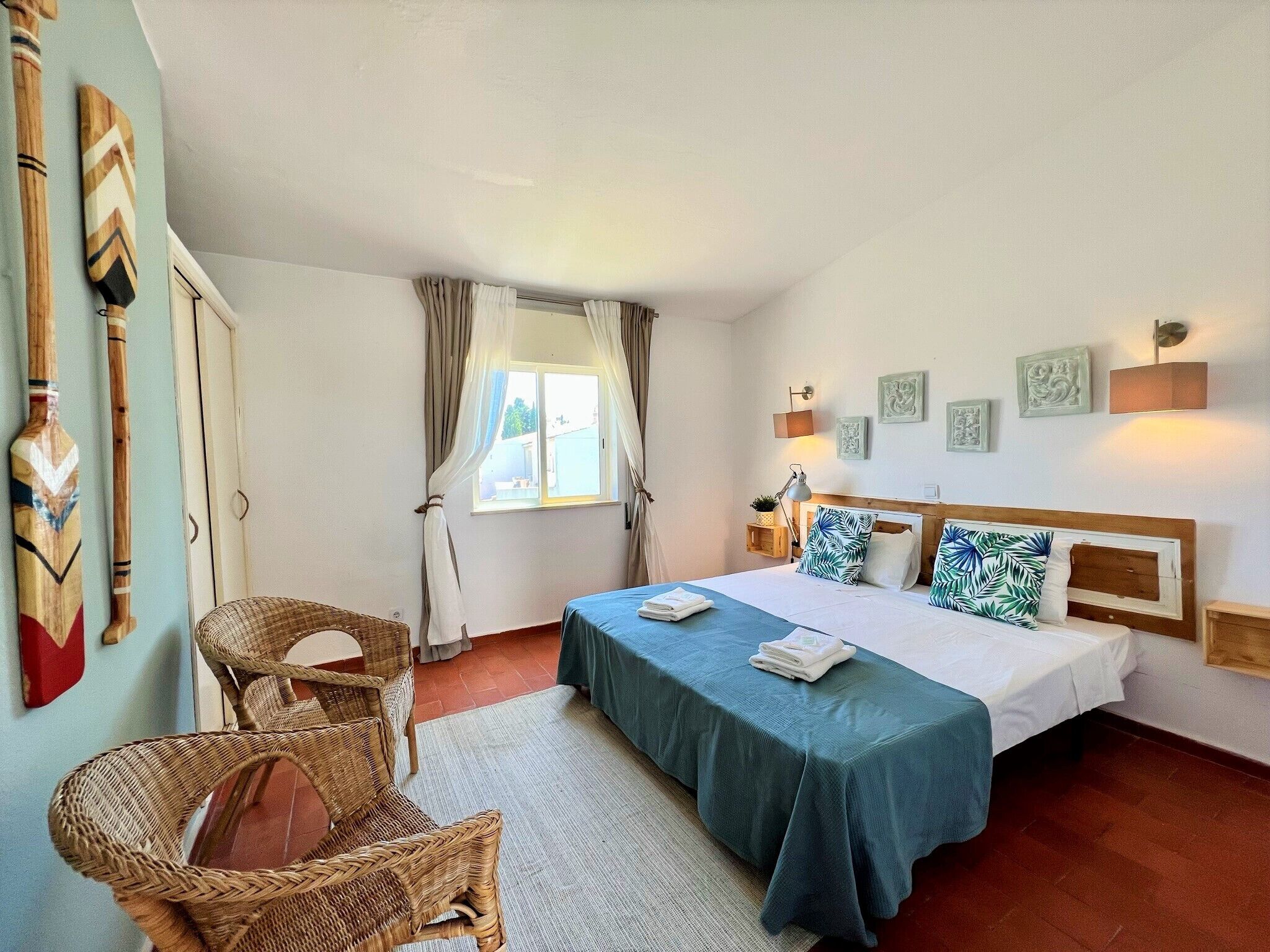 A bedroom with a bed, two wicker chairs, a window with curtains, and wall-mounted lights.