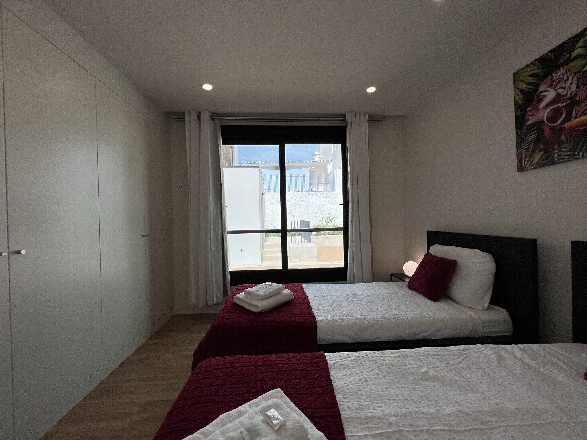 A hotel room with two beds, a large window with curtains, a painting on the wall, and a wardrobe.