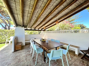 Eine Terrasse im Freien mit einem Holztisch und Stühlen, einer weißen Wand mit Zierleisten und Blick auf Grünflächen und Bäume.