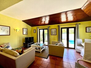 Ein Wohnzimmer mit einer Couch, einem Couchtisch, einem Fernseher und einem Essbereich mit Stühlen. Große Fenster mit Vorhängen, eine Decke mit Holzbalken und ein Fliesenboden sind ebenfalls vorhanden.