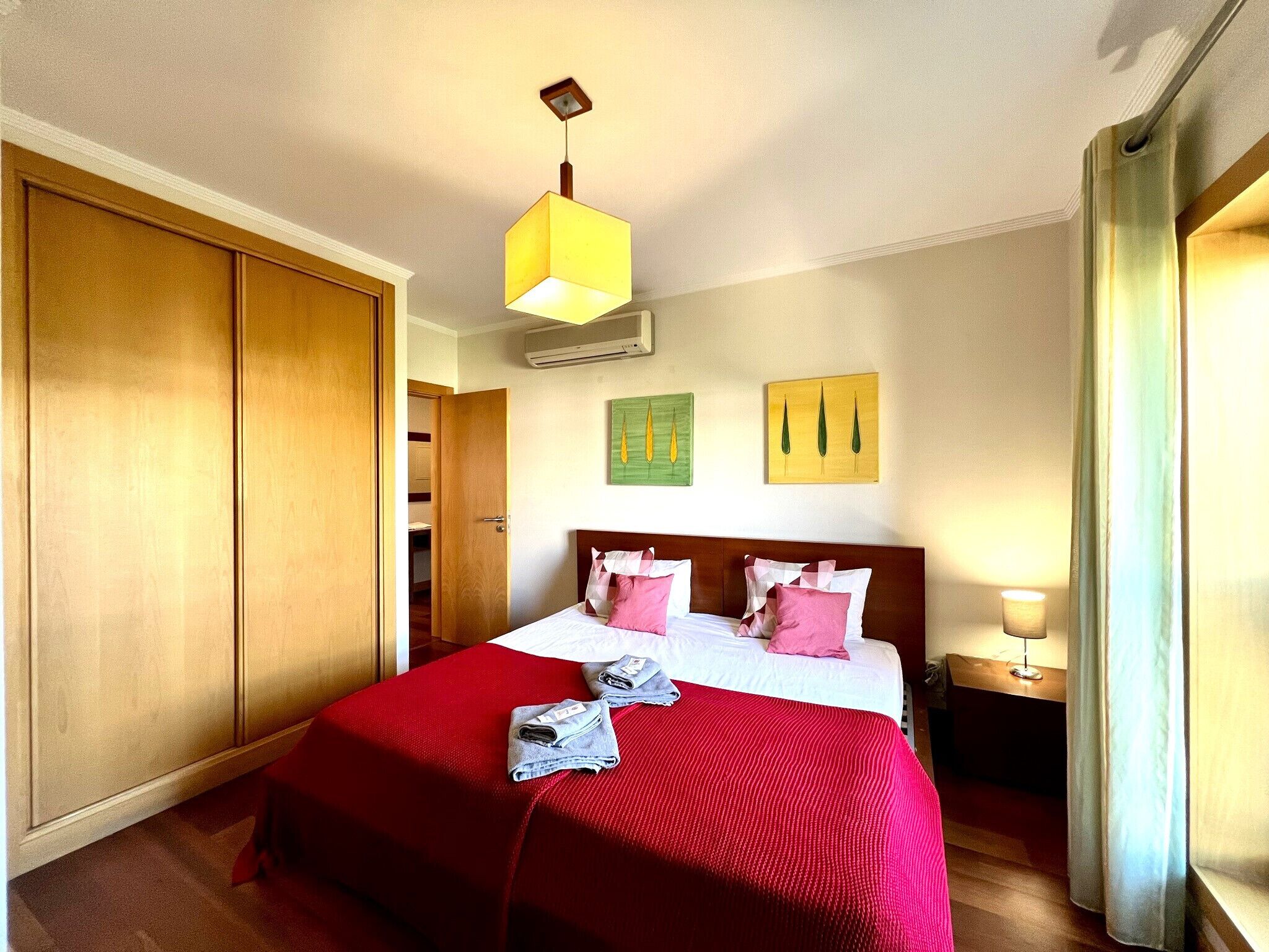 A hotel room with a large bed, a wooden wardrobe, a red bedspread, and two wall-mounted paintings.
