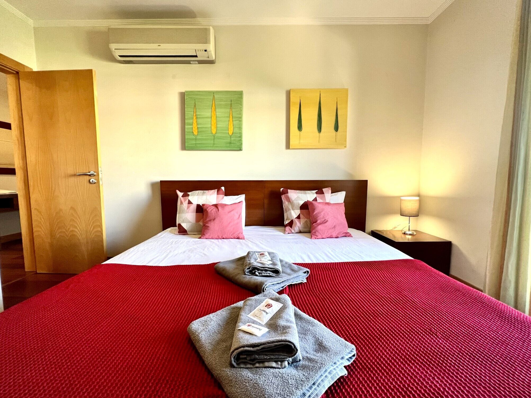 Habitación de hotel con cama, dos almohadas, mesita de noche con lámpara y una obra de arte en la pared.