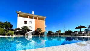 Una piscina con acqua cristallina, circondata da una spiaggia sabbiosa. Sullo sfondo, un edificio a più piani con facciata bianca e arancione.
