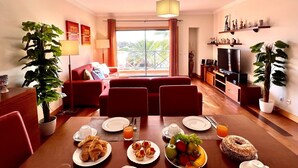 Un soggiorno moderno con divano, tavolo da pranzo e TV. Sono presenti piante, un balcone con vista e un’area pranzo con articoli per la colazione.