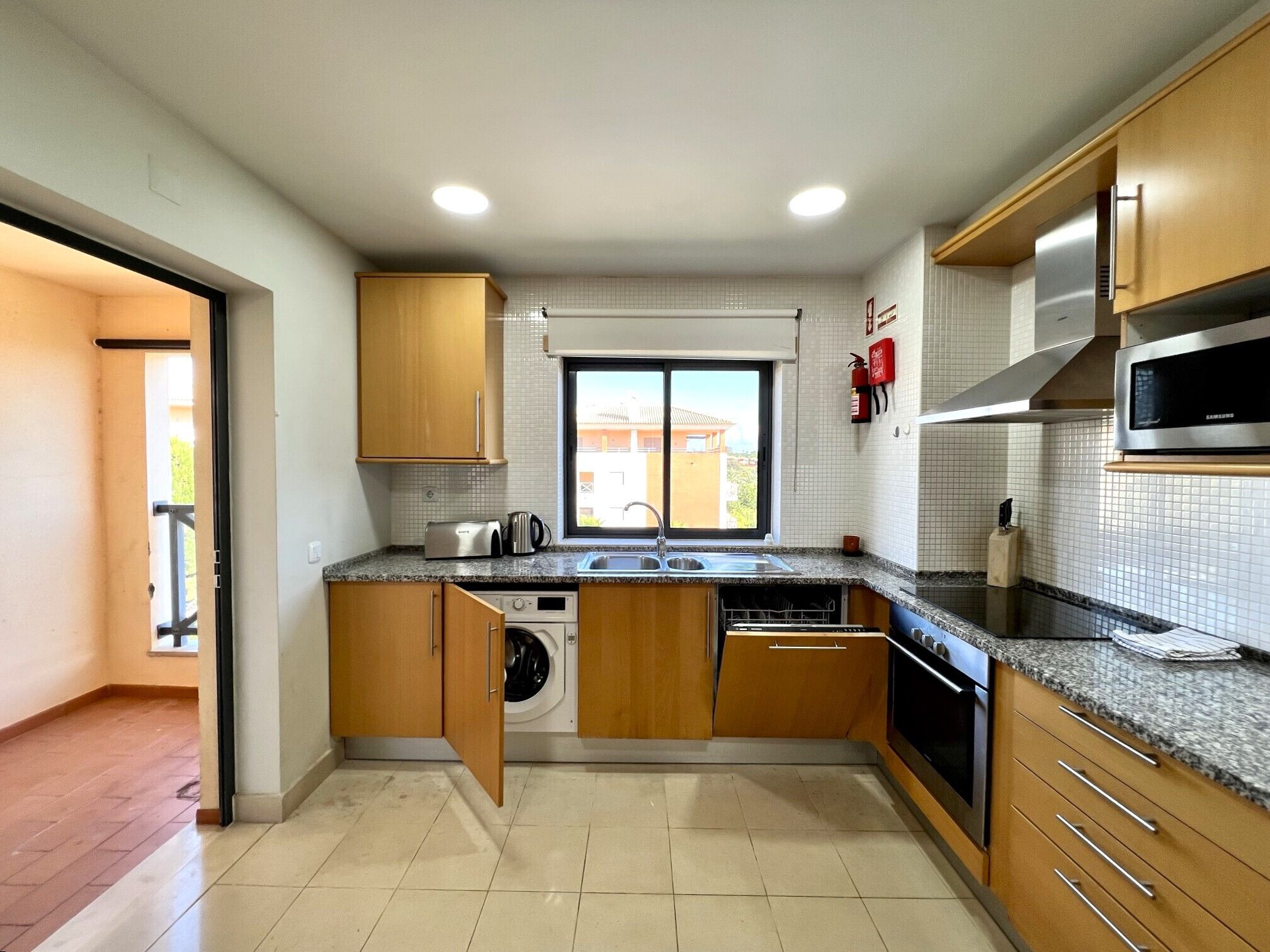 A compact kitchen with wooden cabinets, a built-in microwave, a washing machine, and a window with a view of buildings.