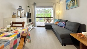 A cozy living room with a sofa, a dining table set for two, a TV, and a large window offering a view of greenery. - Albufeira Cosy by Homing (Albufeira)