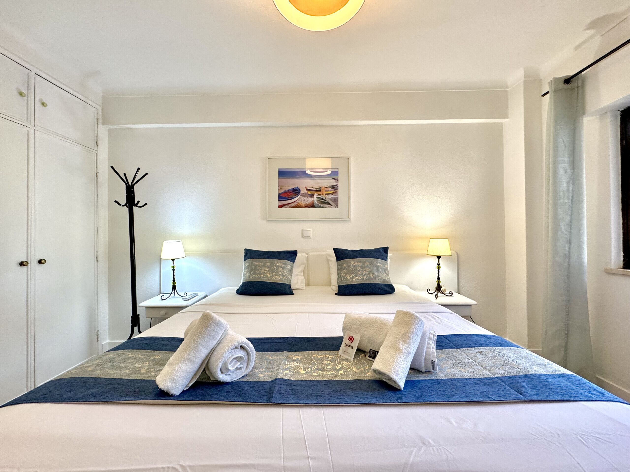 A neatly made bed with a blue and white bedspread, two pillows, and two rolled towels on a white wall with a framed picture above the bed.