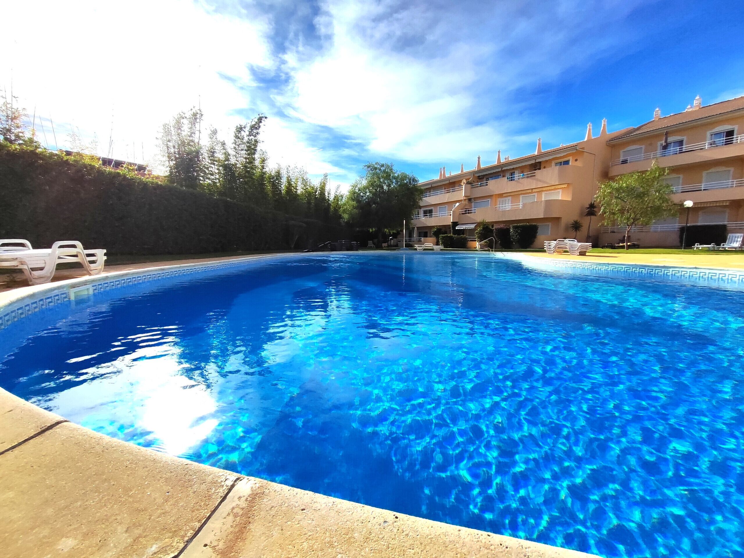 Una piscina con agua azul cristalina, rodeada de sillas de descanso y un edificio con varios balcones.