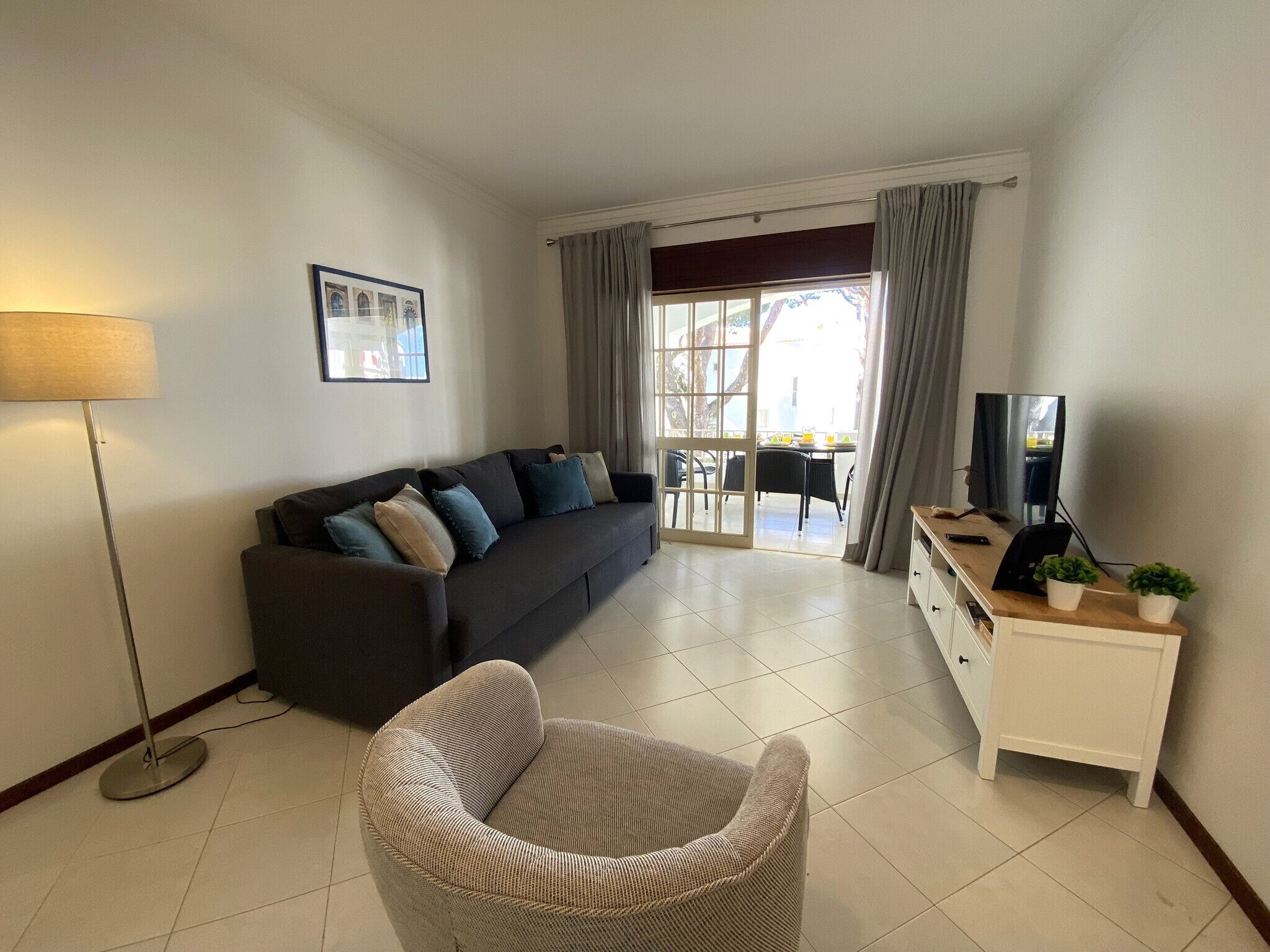 Un salón con sofá, una silla, televisión y una lámpara. Se ve una ventana con cortinas y vistas al exterior.