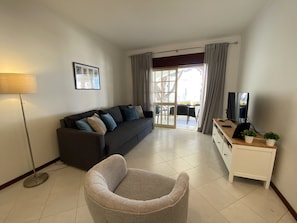 A living room with a sofa, a chair, a TV, and a lamp. There is a window with curtains and a view of the outside. - Vilamoura Garden View 2 With Pool (Loulé)
