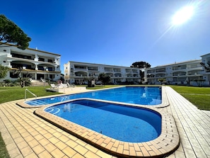 A swimming pool with a tiled deck, surrounded by a building with multiple balconies and a clear sky. - Vilamoura Garden View 2 With Pool (Loulé)