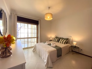 A bedroom with a bed, bedside tables, a mirror, and a window with curtains. - Vilamoura Garden View 2 With Pool (Loulé)