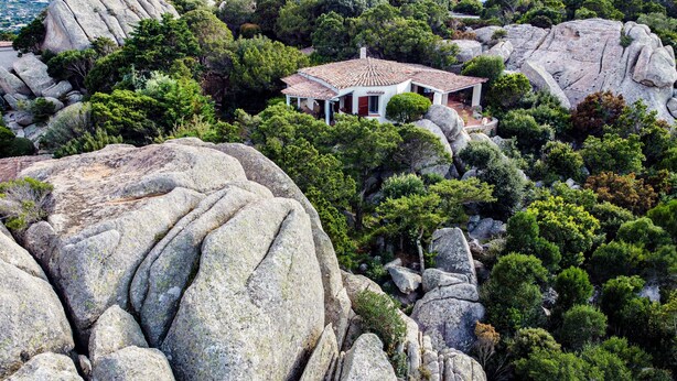 Un edificio blanco con tejado de tejas, rodeado de grandes rocas y abundante vegetación.