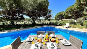 A poolside dining area with a table set for breakfast, surrounded by trees and a clear blue sky. - Vilamoura Amazing Golf Villa With Pool (Loulé)