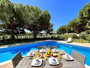 Ein Essbereich am Pool mit gedecktem Tisch für das Frühstück, umgeben von Bäumen und blauem Himmel.