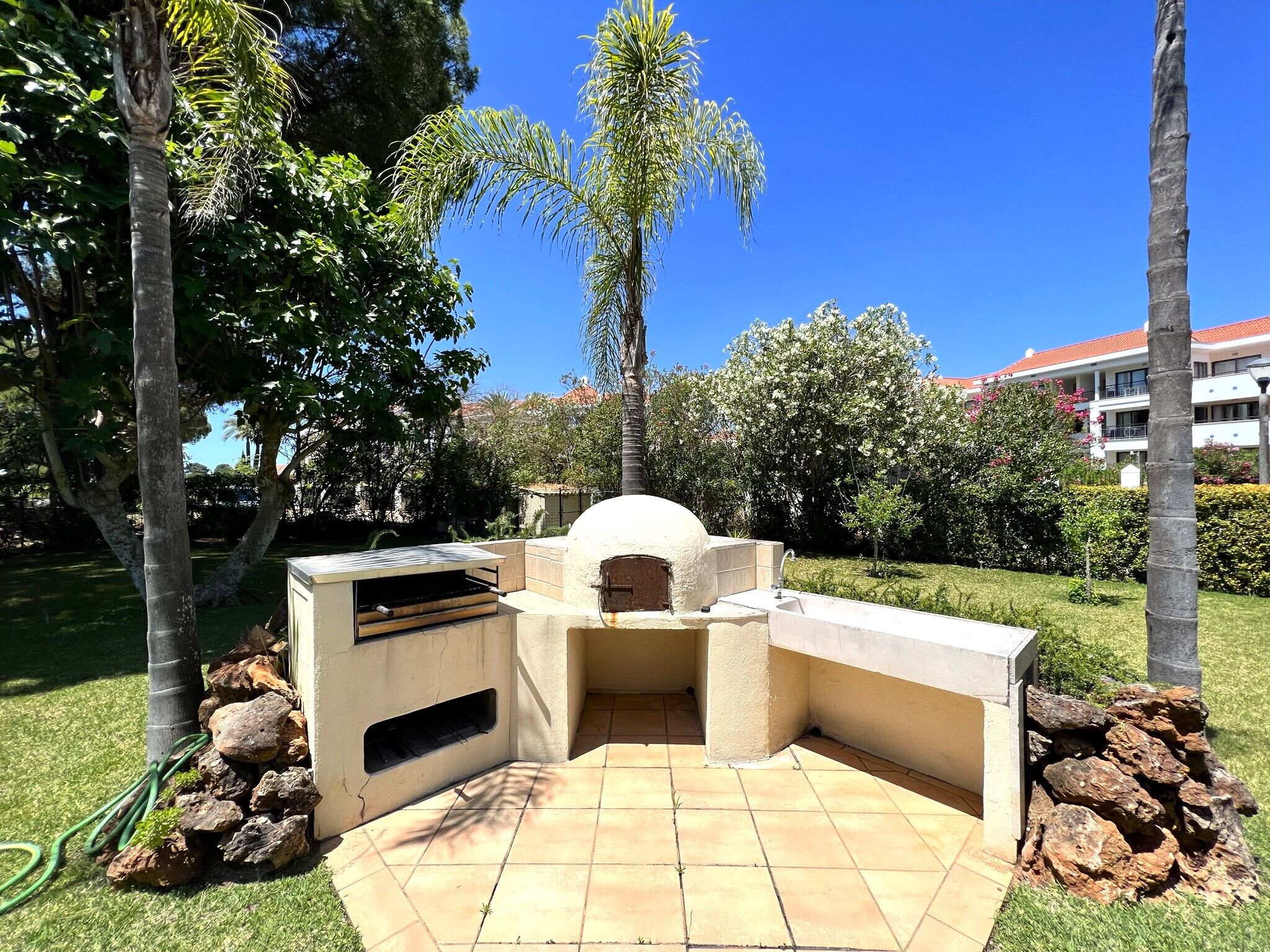 An outdoor stone oven with a tiled patio, surrounded by greenery and palm trees.