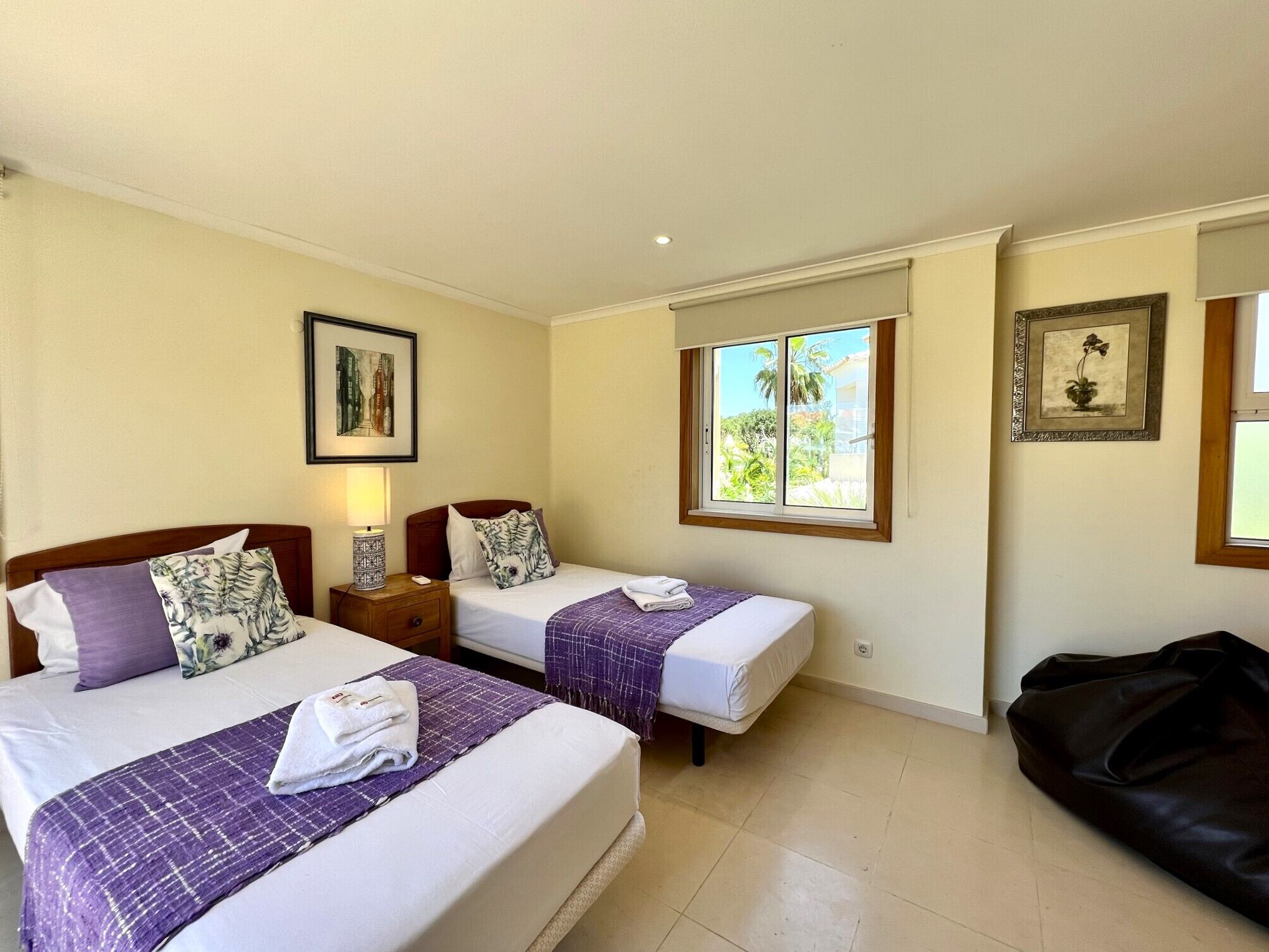A hotel room with two beds, a window with a view of greenery, and a black leather couch.