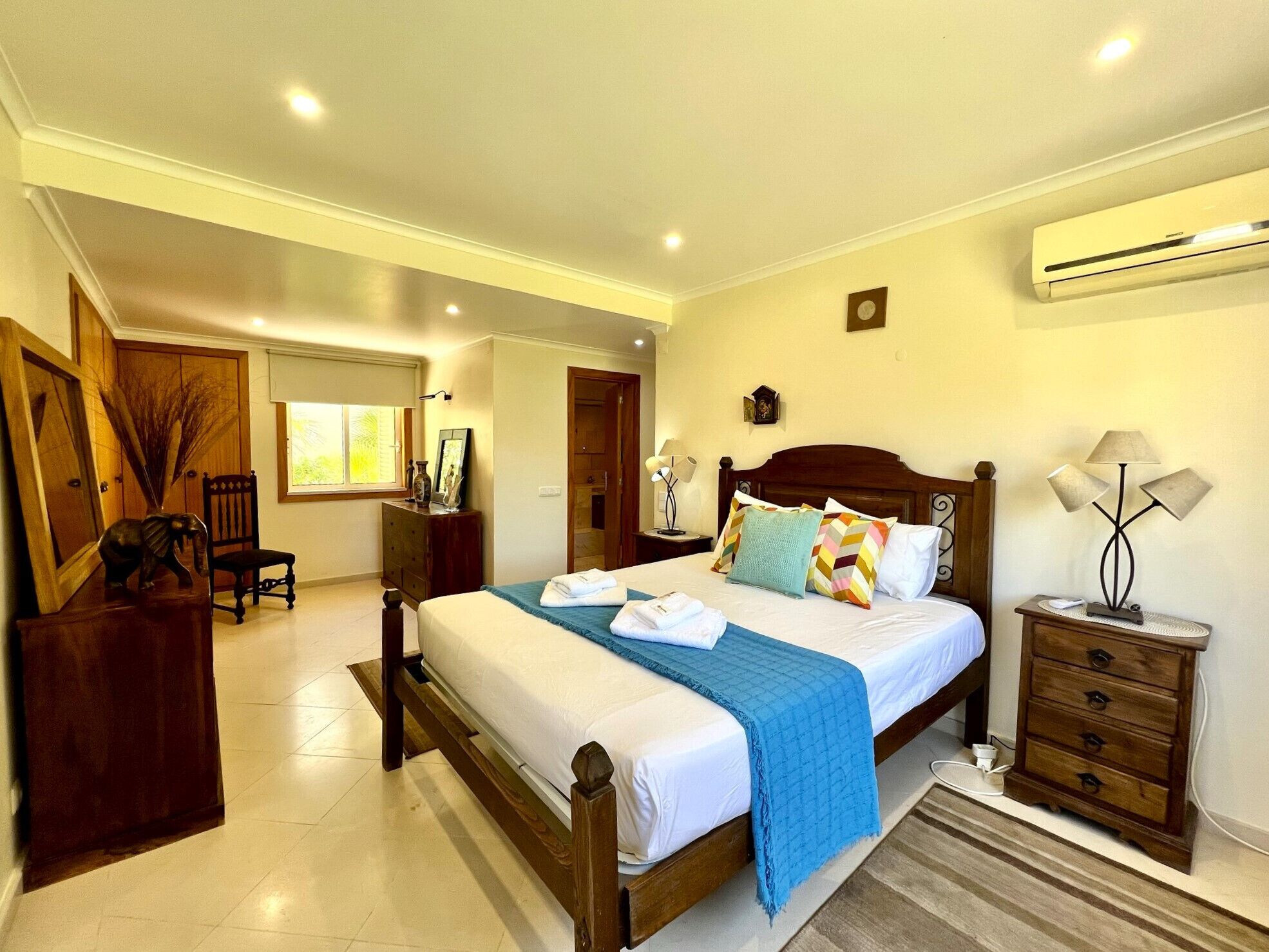 A well-lit bedroom with a large bed, bedside tables, an air conditioning unit, and a view of the outside.