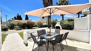 Un patio al aire libre con una mesa redonda y sillas, un parasol grande y una parrilla.