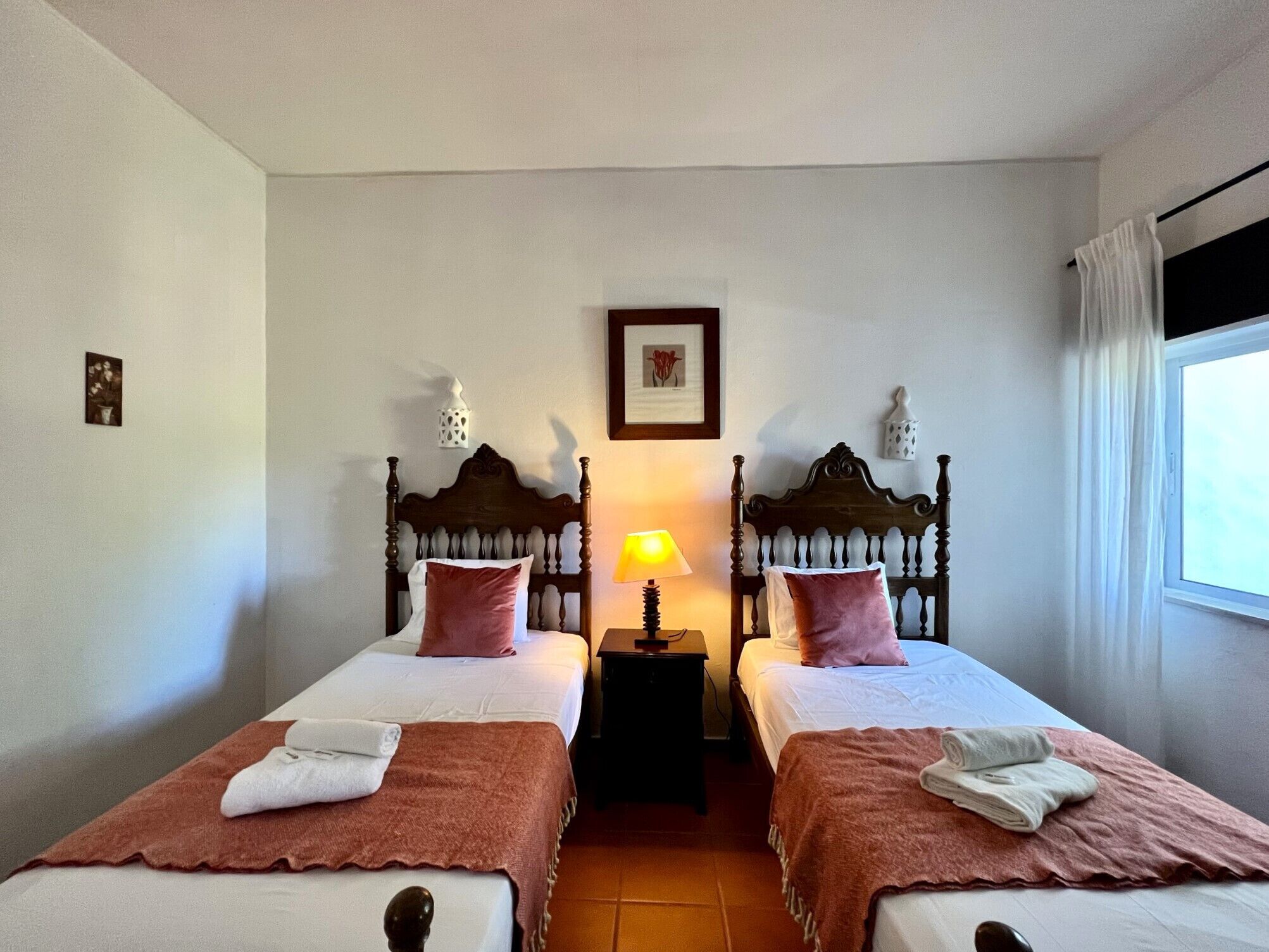 Une chambre d’hôtel avec deux lits simples, une table de chevet avec une lampe, un tableau encadré au mur et une fenêtre avec des rideaux.