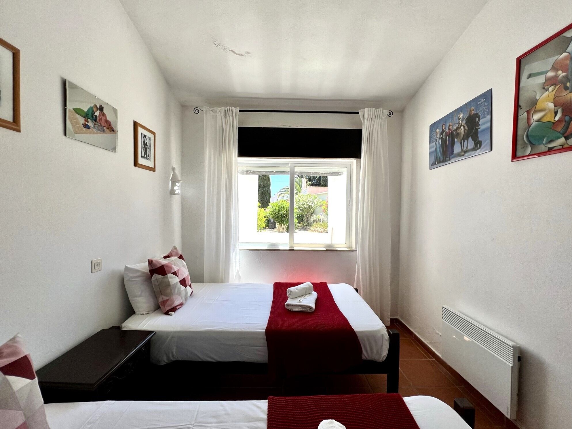 Habitación de hotel con dos camas, una ventana con cortinas, un radiador y cuadros en las paredes.