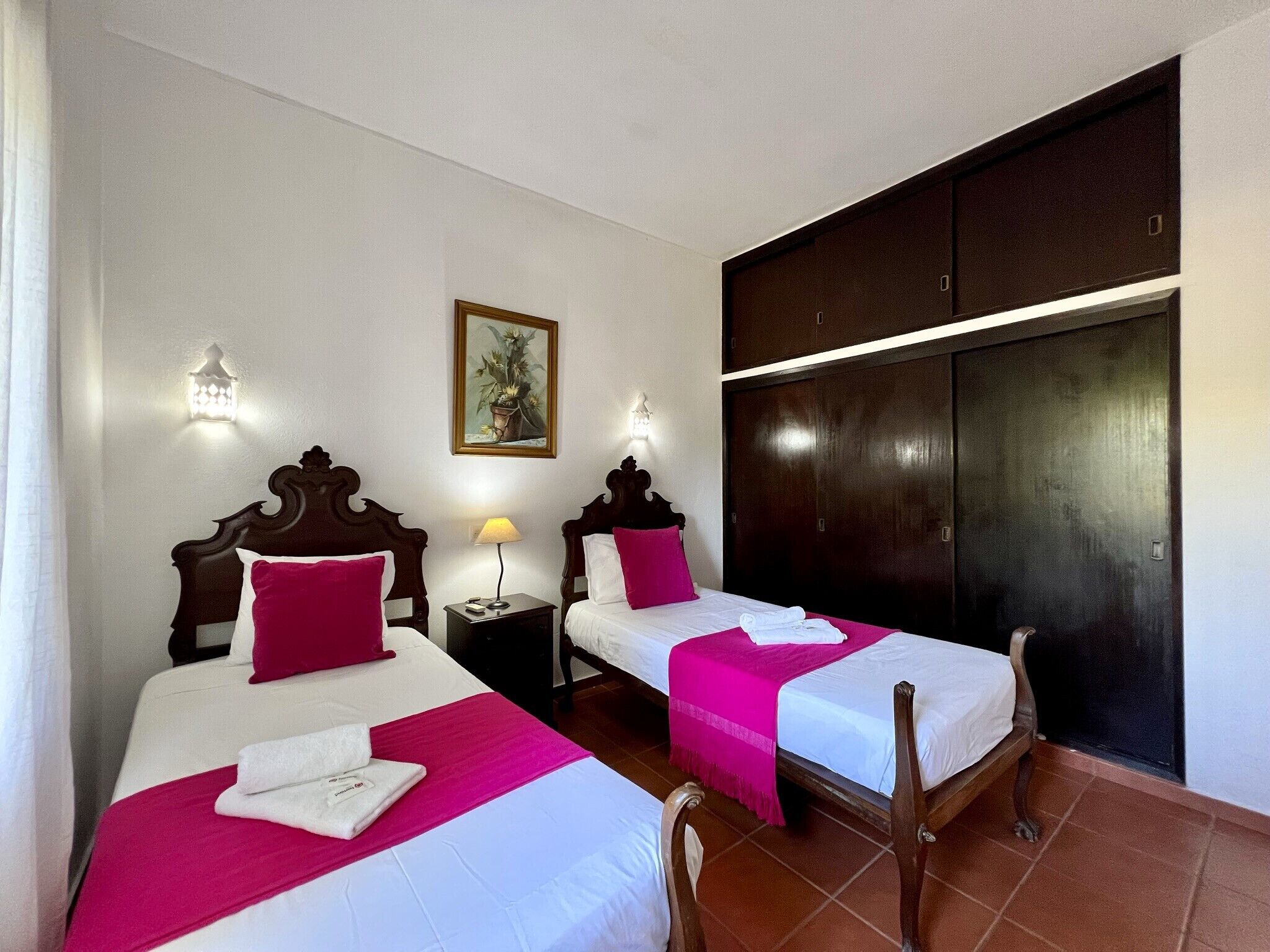 Camera d'albergo con due letti, ciascuno dotato di una testiera in legno, biancheria da letto bianca e tocchi di rosa. Si nota un comodino con una lampada, un quadro appeso alla parete e un armadio con uno specchio.